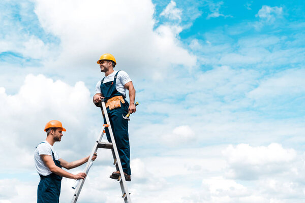 cheerful repairmen standing on ladder and smiling against blue sky with clouds 