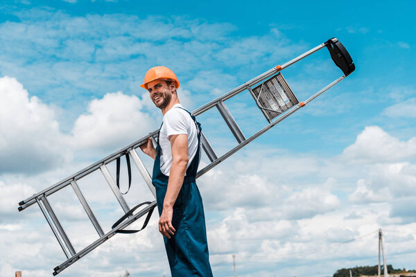 positive repairman holding ladder and smiling against blue sky with clouds 