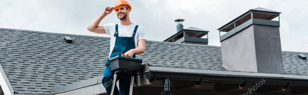 panoramic shot of happy repairman sitting on roof and holding toolbox 