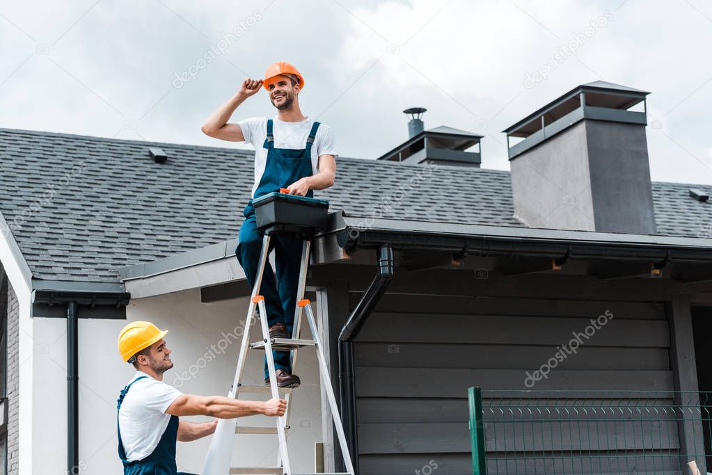 happy repairman sitting on roof and holding toolbox near coworker in helmet