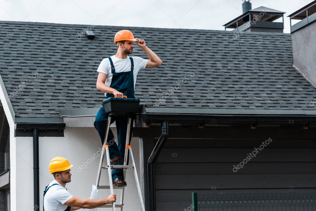 handsome repairman sitting on roof and holding toolbox near coworker in helmet 