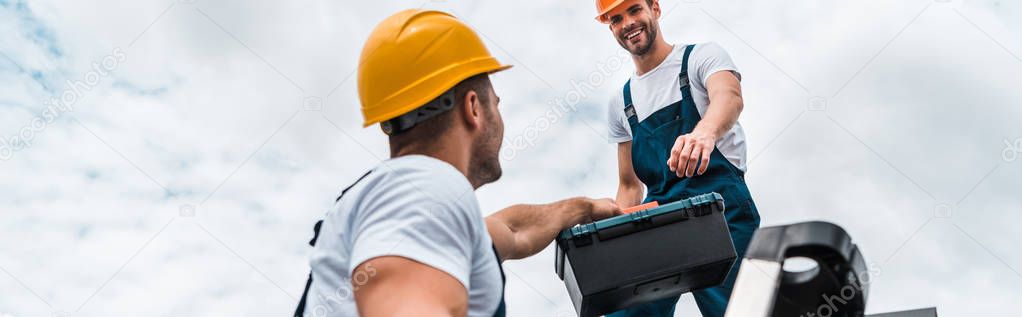 panoramic shot of handyman giving toolbox to happy colleague 