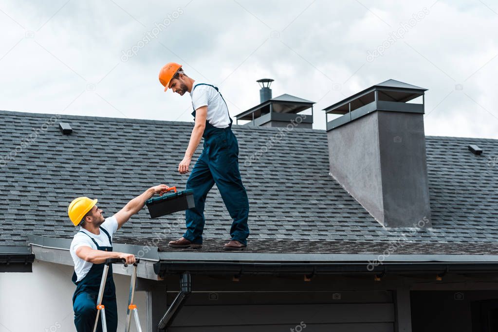 handyman in helmet giving toolbox to handsome colleague standing on rooftop 