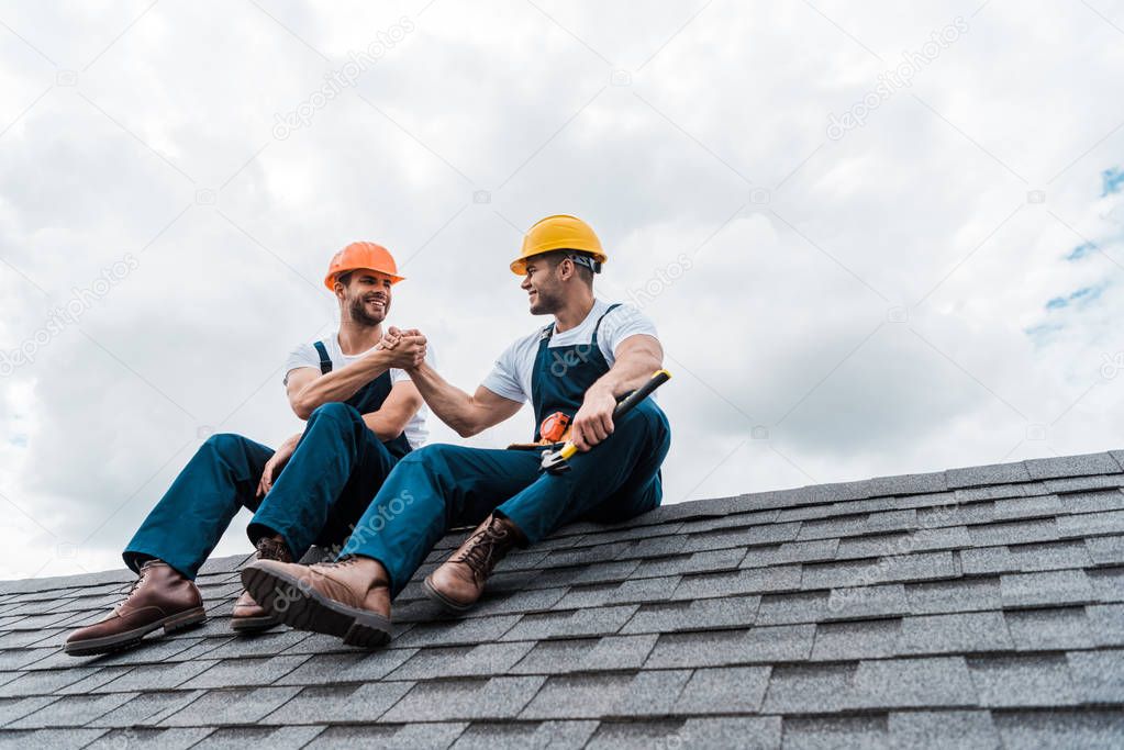 happy handymen in helmets holding hands while sitting on rooftop 