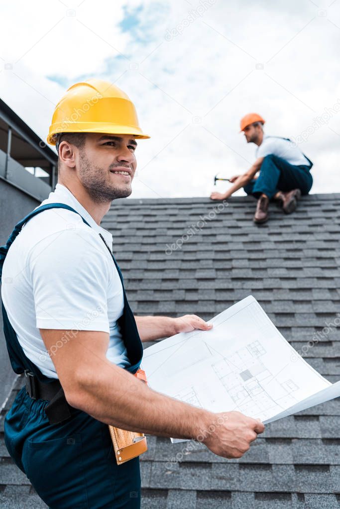 selective focus of handsome builder in helmet holding blueprint near colleague repairing roof 