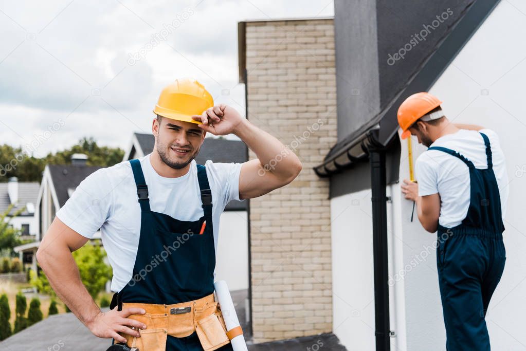 selective focus of cheerful repairman touching helmet and smiling near coworker 