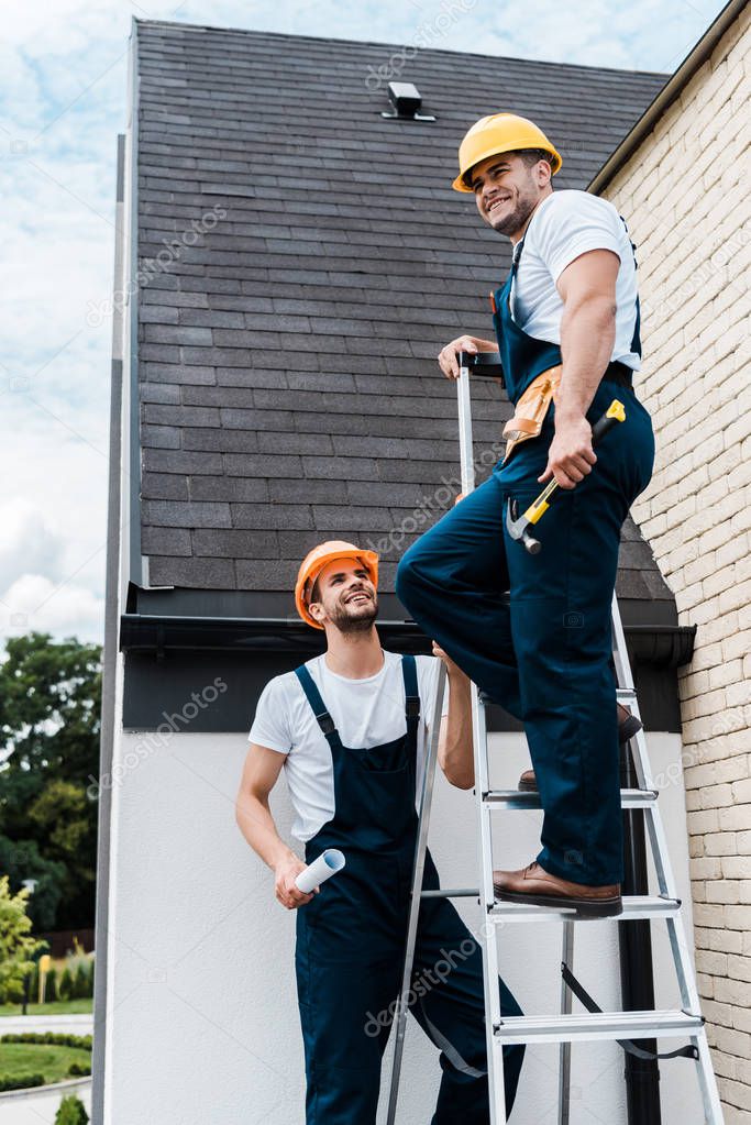 handsome man in uniform holding paper roll and looking at coworker standing on ladder 