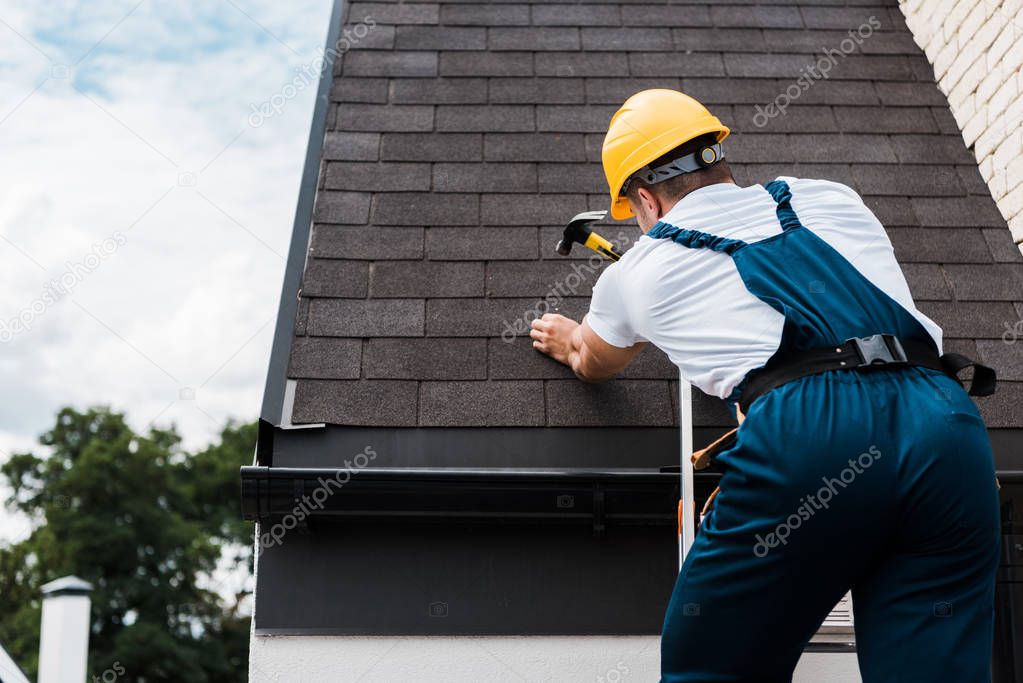 back view of handyman in uniform and helmet repairing roof while standing on ladder 