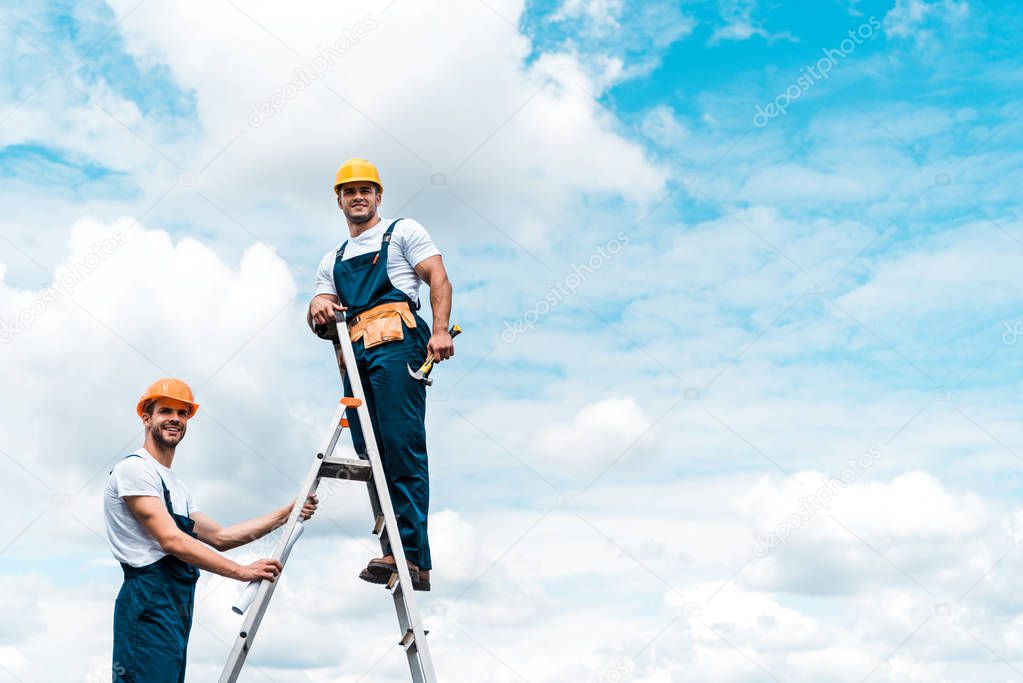 happy repairmen standing on ladder and smiling against blue sky with clouds 
