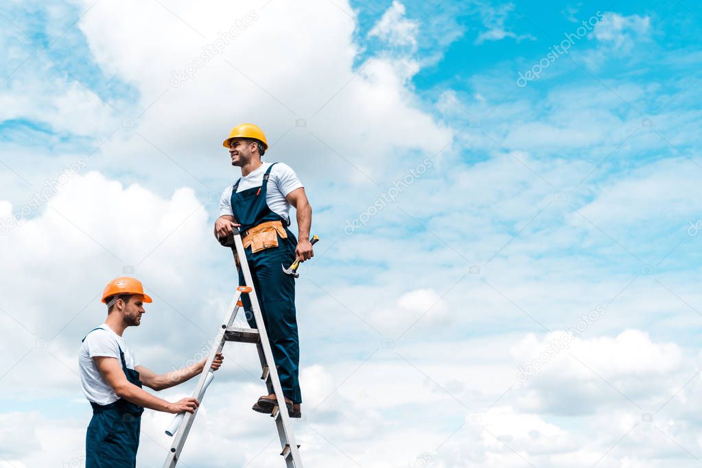 cheerful repairmen standing on ladder and smiling against blue sky with clouds 