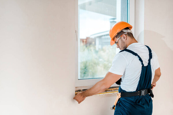 handsome repairman in goggles measuring wall near window 