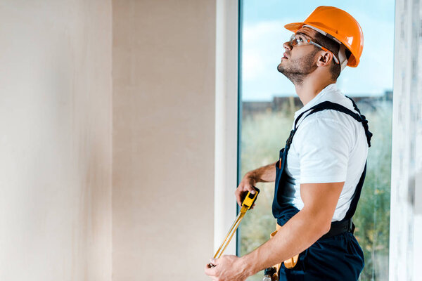 handsome repairman in goggles holding measuring tape and looking at wall near window 