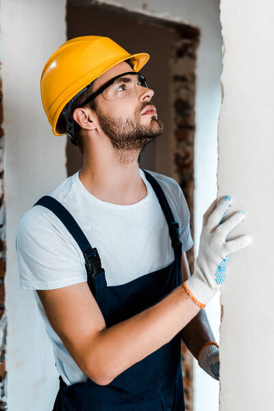 handsome bearded handyman in goggles and helmet looking at wall 