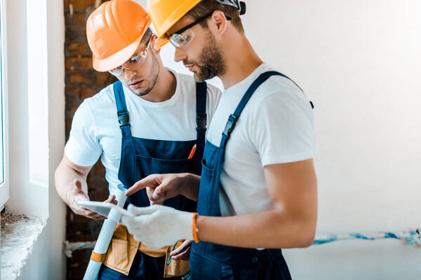 handsome repairman in goggles looking at digital tablet near coworker 