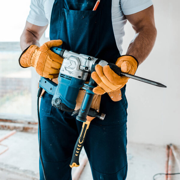 cropped view of workman in uniform and yellow gloves holding hammer drill 