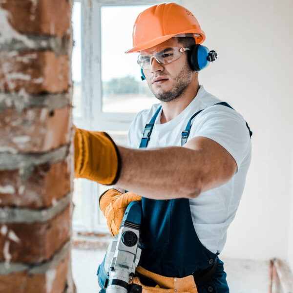 selective focus of workman in uniform and goggles  holding hammer drill and looking at brick wall 