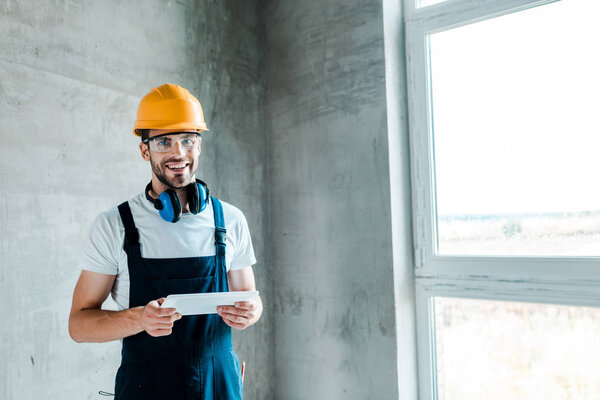 cheerful repairman in helmet and goggles holding digital tablet 