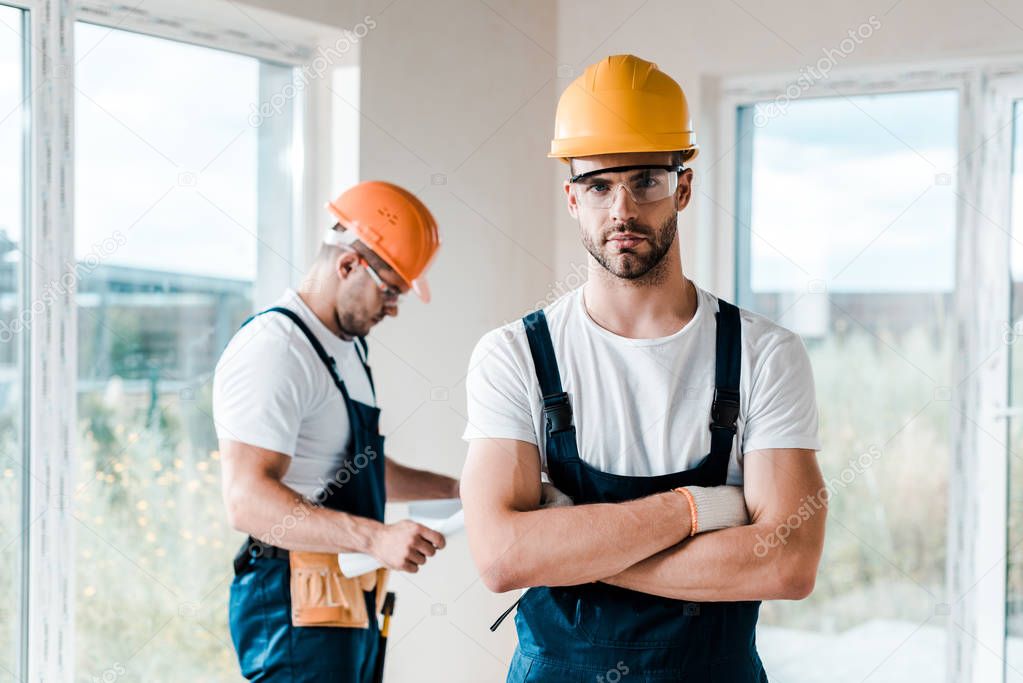selective focus of handsome handyman in goggles standing with crossed arms near coworker 