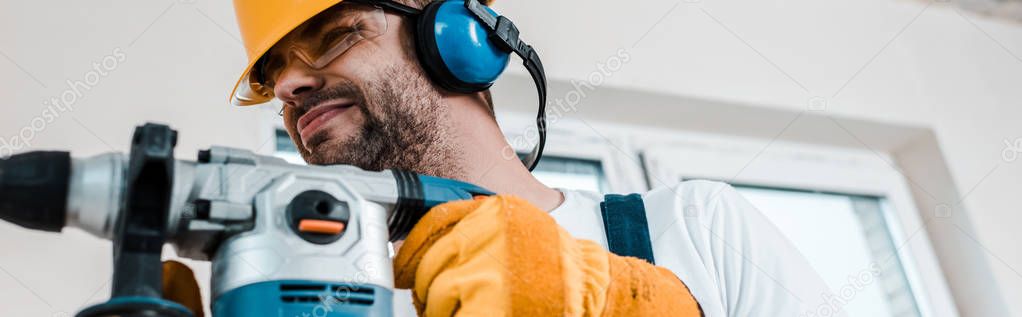 panoramic shot of handyman in helmet using hammer drill 