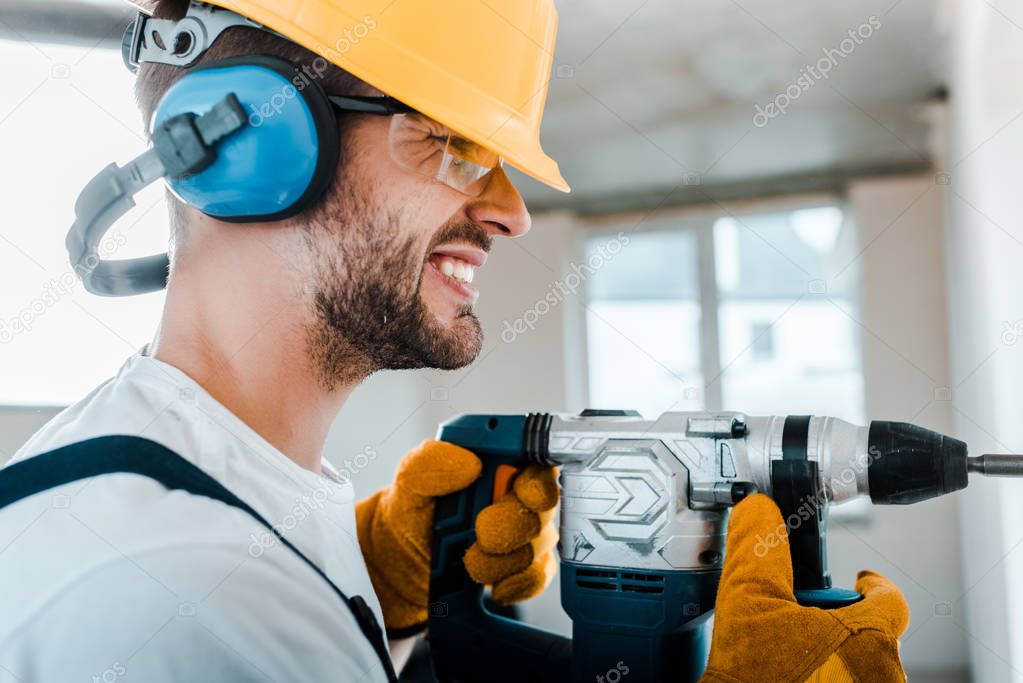 handsome handyman in uniform and yellow gloves using hammer drill 