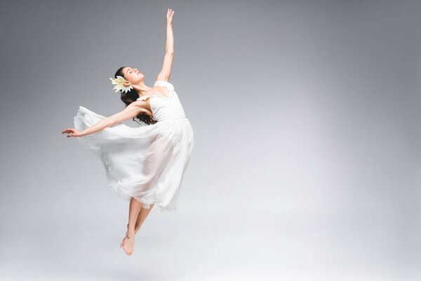 graceful ballerina in white dress jumping while dancing on grey background