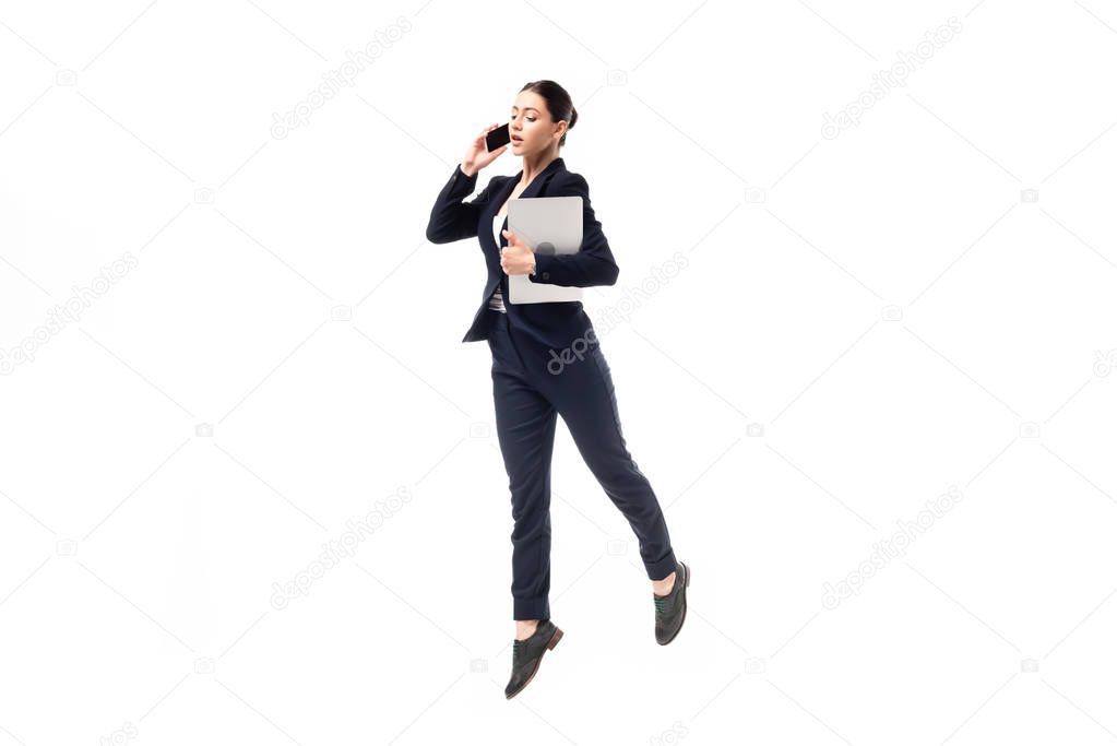 young businesswoman talking on smartphone and holding laptop while dancing isolated on white