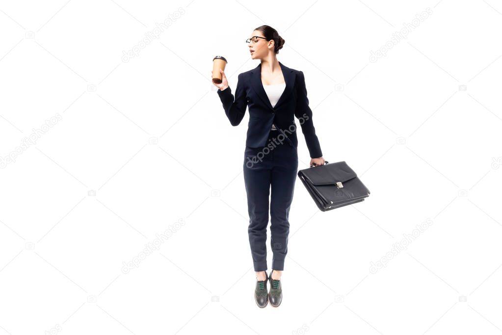 young businesswoman holding coffee to go and briefcase while jumping isolated on white