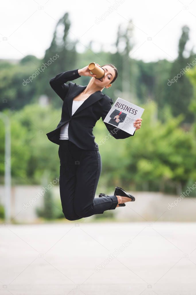elegant businesswoman holding newspaper and drinking coffee to go while levitating on street