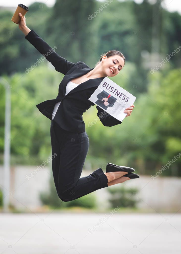 cheerful businesswoman levitating on street while holding coffee to go and business newspaper
