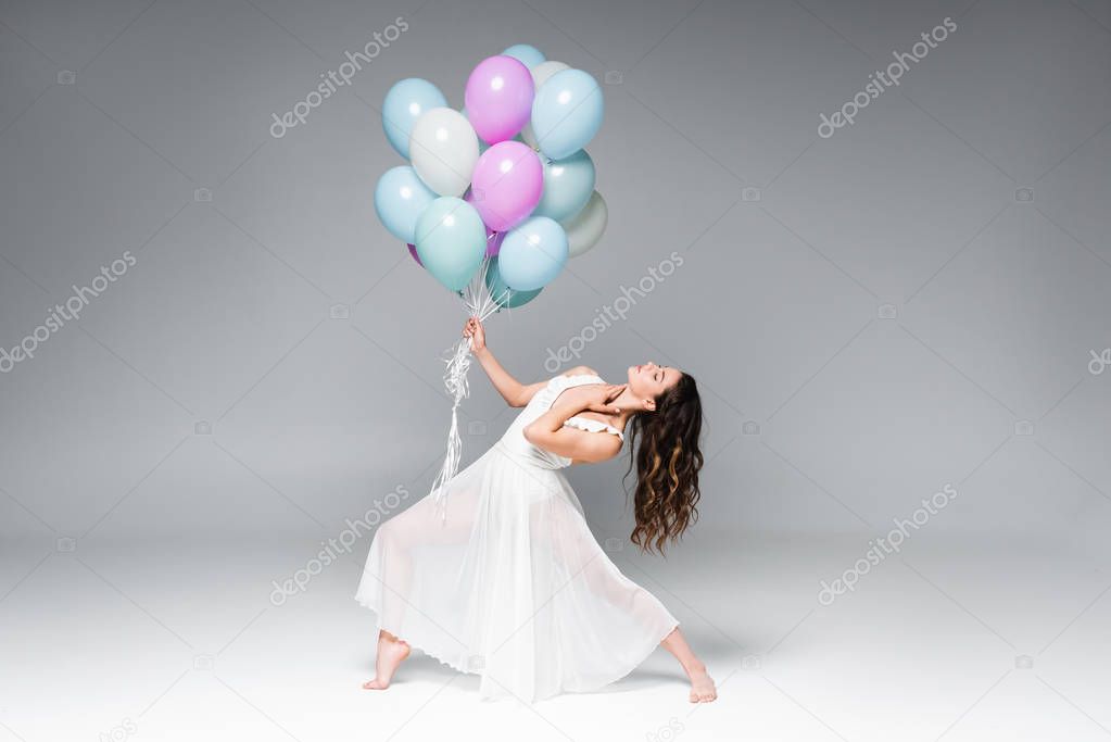 young beautiful ballerina in white dress dancing with festive balloons on grey background