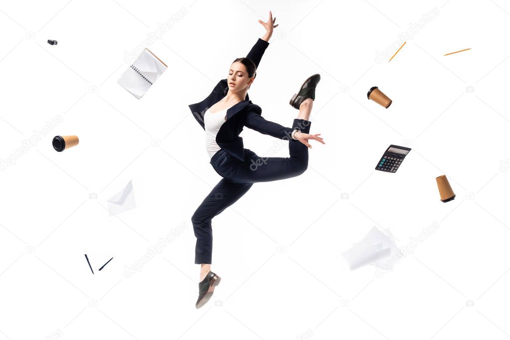 young businesswoman dancing surrounded with paper cups, notebook, papers, calculator, pens and pencil fying around isolated on white