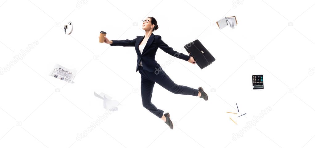 panoramic shot of businesswoman holding coffee to go and briefcase while levitating surrounded headphones, newspaper, calculator and stationery isolated on white