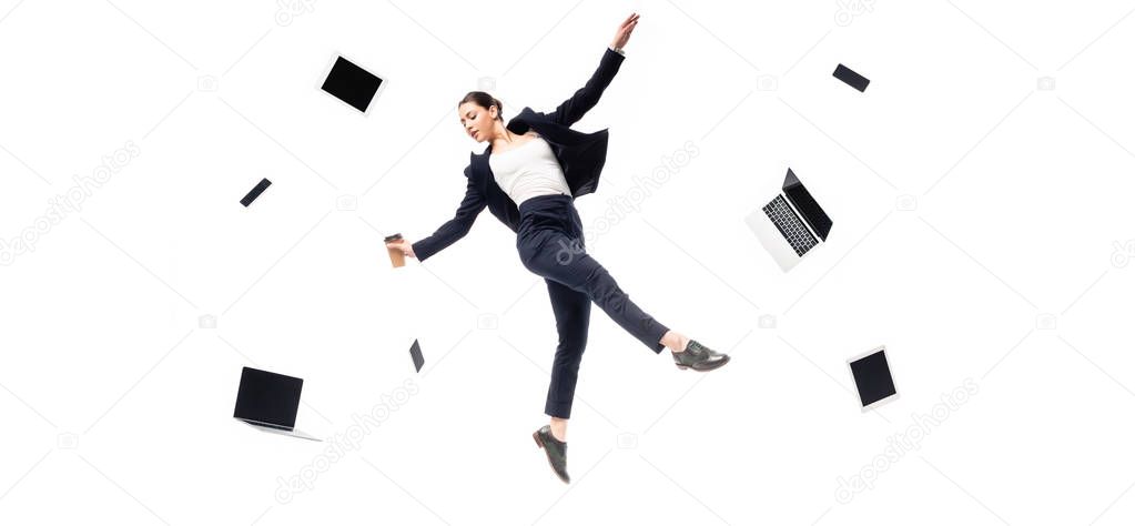 panoramic shot of businesswoman holding paper cup while levitating surrounded with flying laptops and smartphones isolated on white