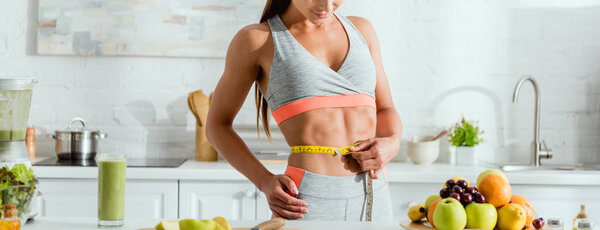 panoramic shot of sportive woman measuring waist near fruits 