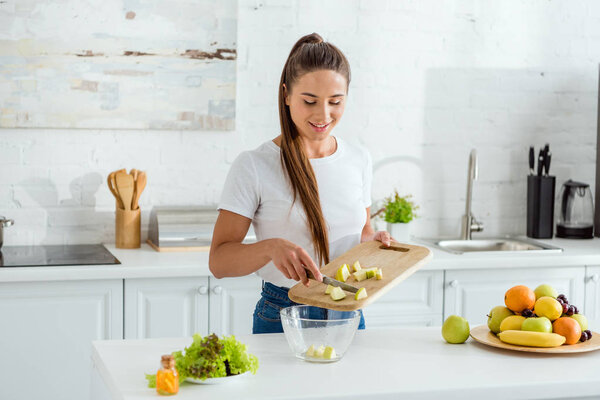 happy young woman putting sliced green apple in bowl 