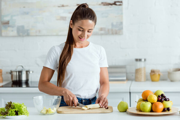 cheerful girl cutting tasty banana on cutting board near fruits 