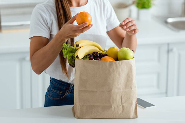 cropped view of woman with orange near paper bag and smartphone with blank screen
