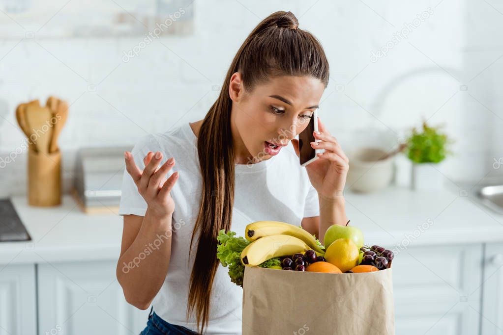 attractive young woman looking at paper bag and gesturing while talking on smartphone 