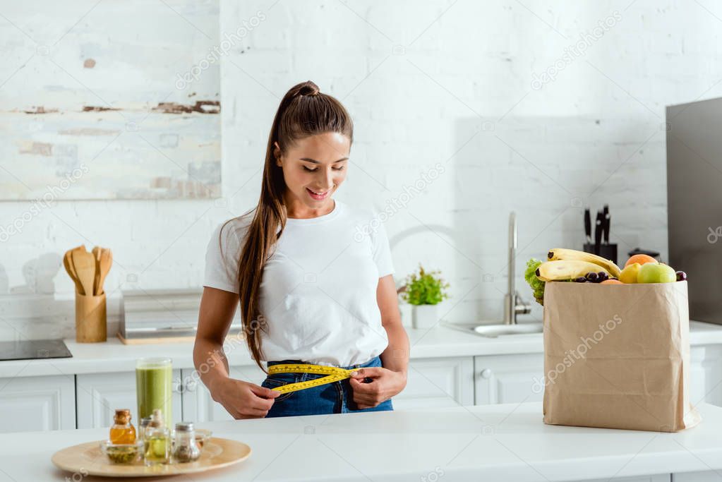 happy young woman measuring waist near groceries 