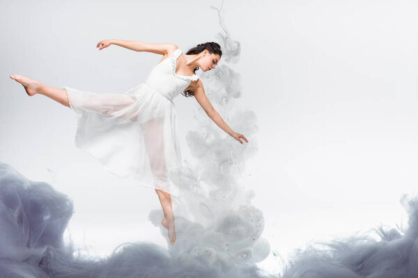 young graceful ballerina in white dress dancing in grey smoke on grey background