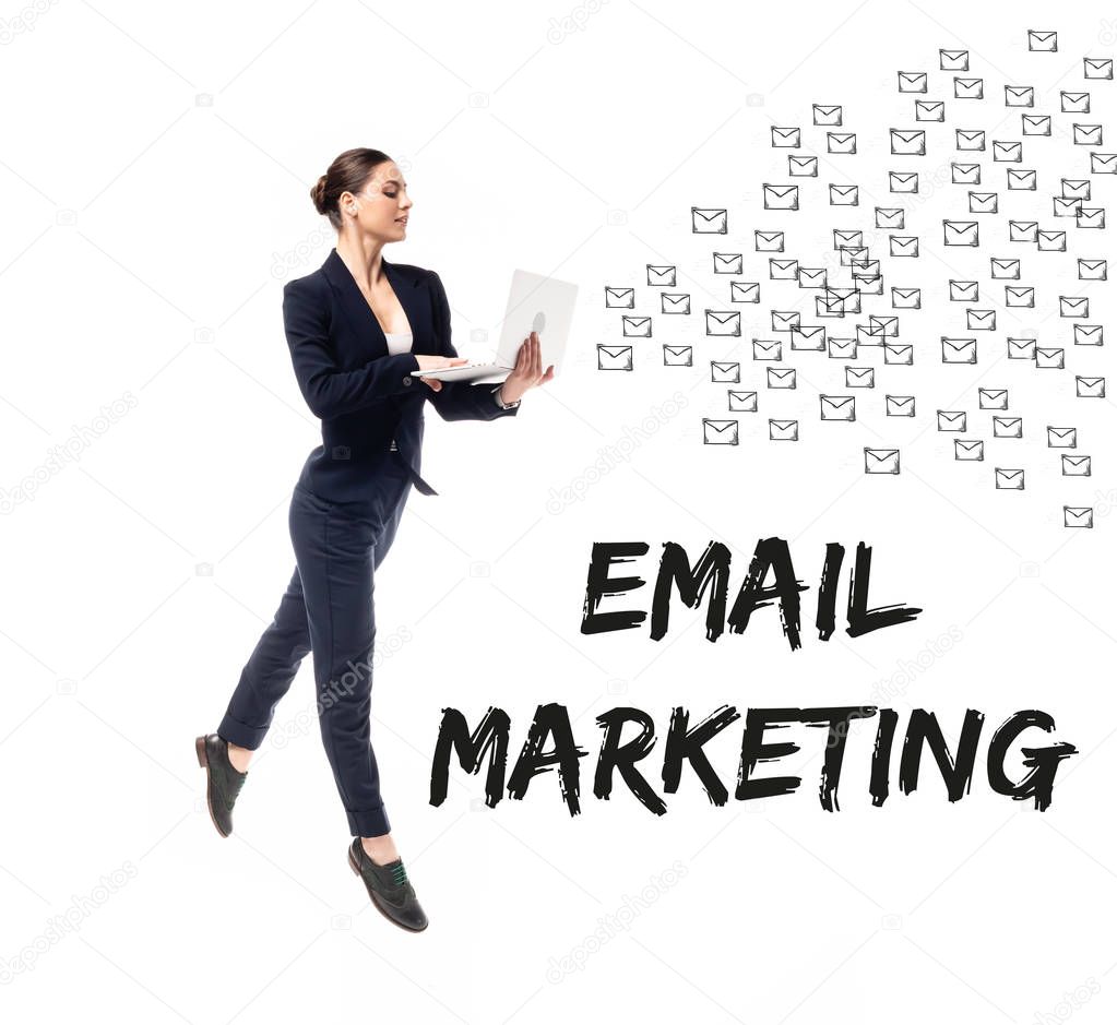 Attractive businesswoman using laptop near email marketing lettering and e-mail icons isolated on white