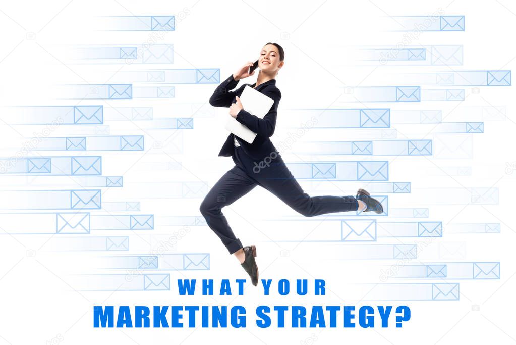 Smiling businesswoman talking on smartphone and holding laptop while levitating on background with e-mail icons and what your marketing strategy question inscription isolated on white