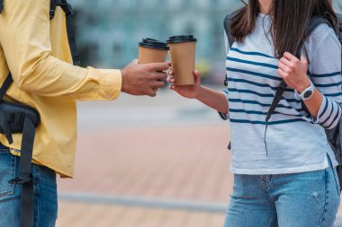 Cropped view of bi-facial man and woman with backpack holding paper cups of coffee at the street  clipart