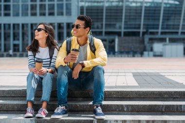 Sırt çantaları ile ırklar arası turistler kahve kağıt bardak ile merdivenlerde oturan ve uzağa bakıyor