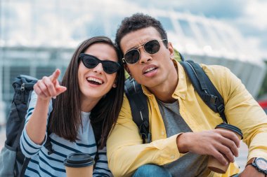 Kadın parmak ile işaret ve sırt çantası ve kahve fincanı ile ilgilenen iki ırklı arkadaş yakınında gülümseyen