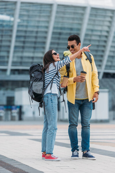 Woman pointing with finger near bi-racial man with coffee paper cup and backpack