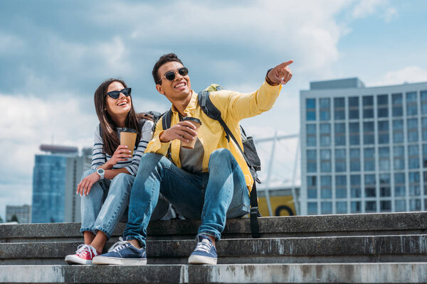 Mixed race tourist with coffee cup sitting on stairs near woman and pointing with finger