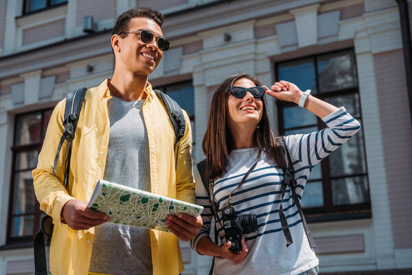 Woman with digital camera sightseeing near bi-racial friend with map and backpack