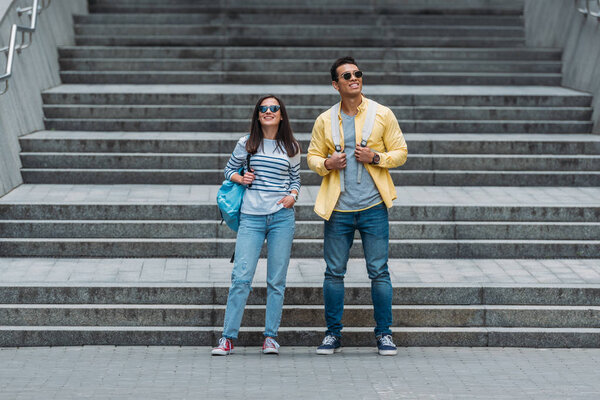 Smiling woman standing in front of stairs near bi-racial friend with backpack