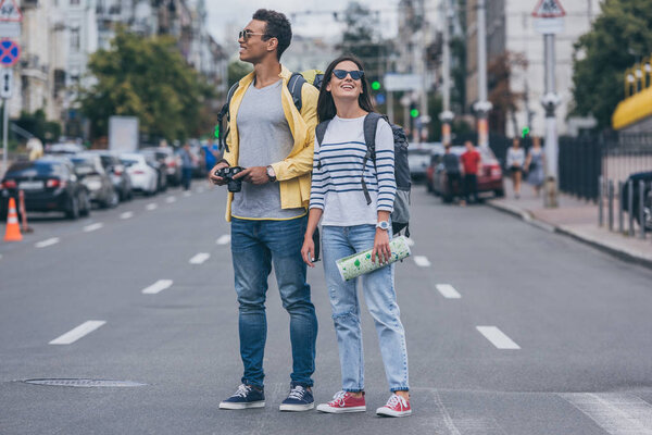 Woman holding map and standing on road near bi-racial friend with backpack and digital camera